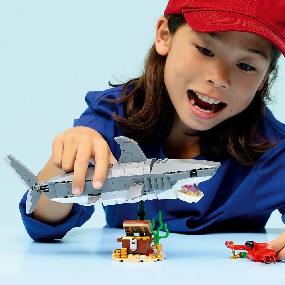 LEGO Creator shark toy in gray plastic bricks as a child holds and plays with the shark above a small treasure chest scene, emphasizing scale, angular shapes, and imaginative indoor ocean play.