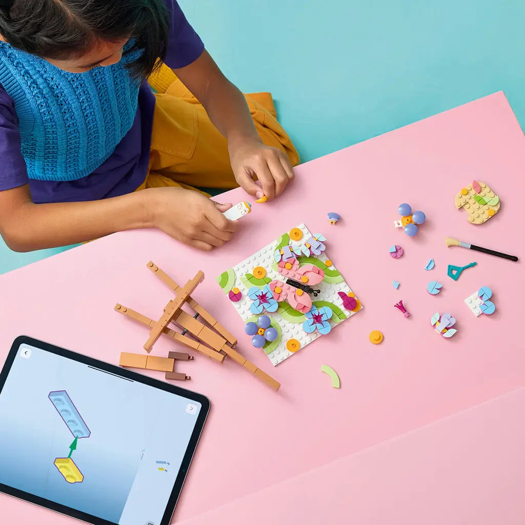 LEGO Creator Floral Art in pink, blue, green and purple plastic, as a child assembles a butterfly flower painting beside loose pieces, easel and tablet on a pink table.