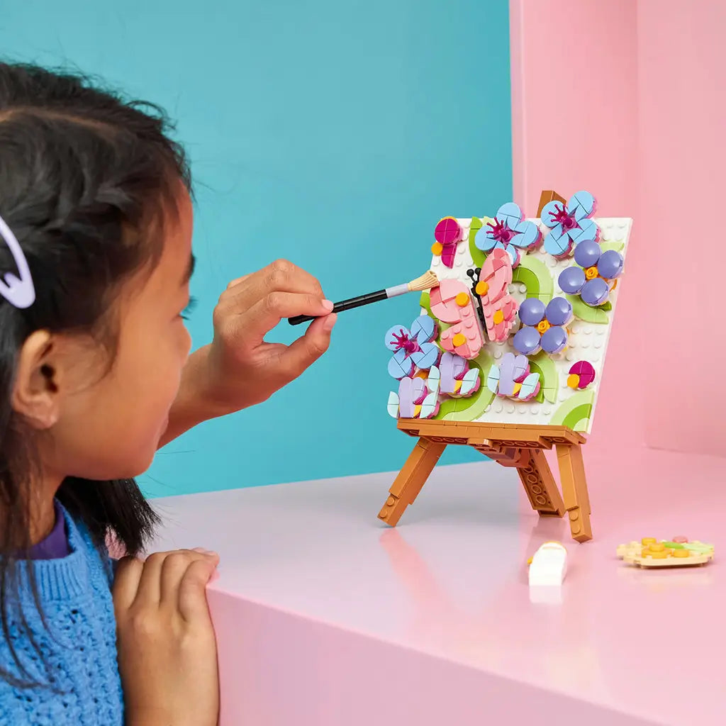LEGO Creator Floral Art in pink, blue, green and purple plastic, as a child uses a brush beside a butterfly and flower painting displayed on a brown brick-built easel.