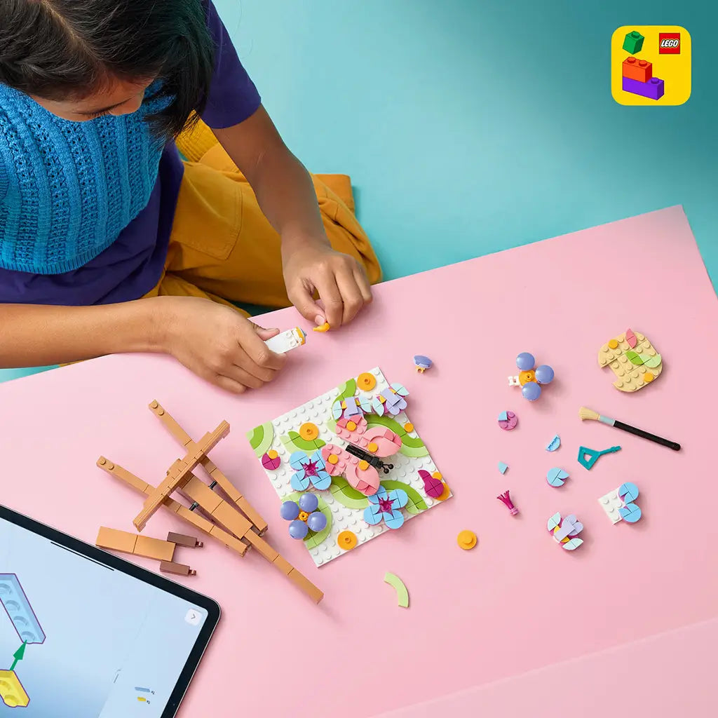 LEGO Creator Floral Art in pink, blue, green and purple plastic, as a child builds a butterfly flower painting among loose pieces, easel, brush and tablet on a pink table.