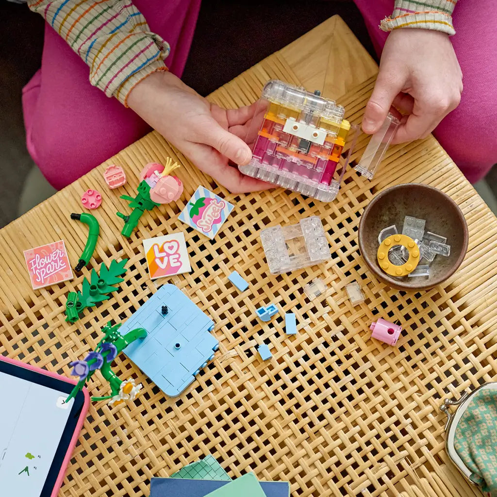 LEGO Creator floral decor in pink, clear, green, and blue plastic bricks is assembled by a child on a woven table with flower pieces, tiles, and a tablet nearby.