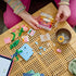 LEGO Creator floral decor in pink, clear, green, and blue plastic bricks is assembled by a child on a woven table with flower pieces, tiles, and a tablet nearby.