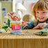 LEGO Creator floral perfume bottle in clear, pink, yellow, green, and blue plastic bricks sits on a woven table as a smiling child lifts its transparent stopper.
