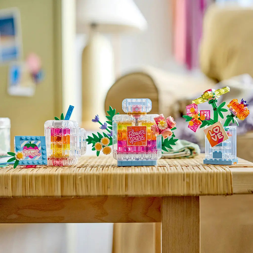 LEGO Creator floral decor builds in clear, pink, yellow, green, and blue plastic bricks display a perfume bottle, compact, frame, and flower vase on a woven table.