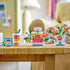 LEGO Creator floral decor builds in clear, pink, yellow, green, and blue plastic bricks display a perfume bottle, compact, frame, and flower vase on a woven table.