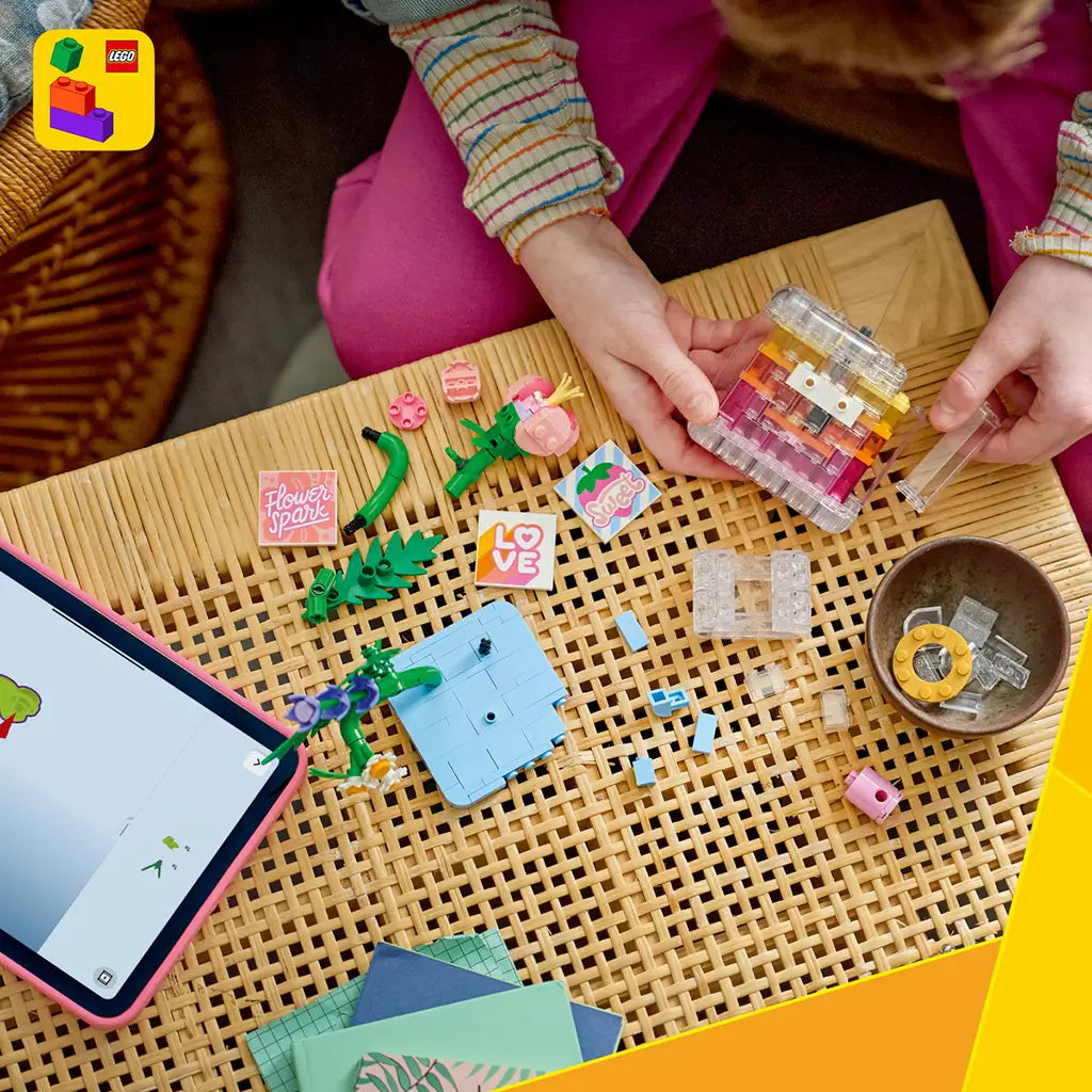 LEGO Creator floral decor set in pink, clear, green, and blue plastic bricks shows a child building a perfume bottle and flowers on a woven table beside a tablet.
