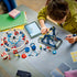 LEGO Creator Ferris Wheel in white, blue and red plastic, as a child assembles the dark blue support frame beside the wheel, loose pieces, minifigures and a tablet.