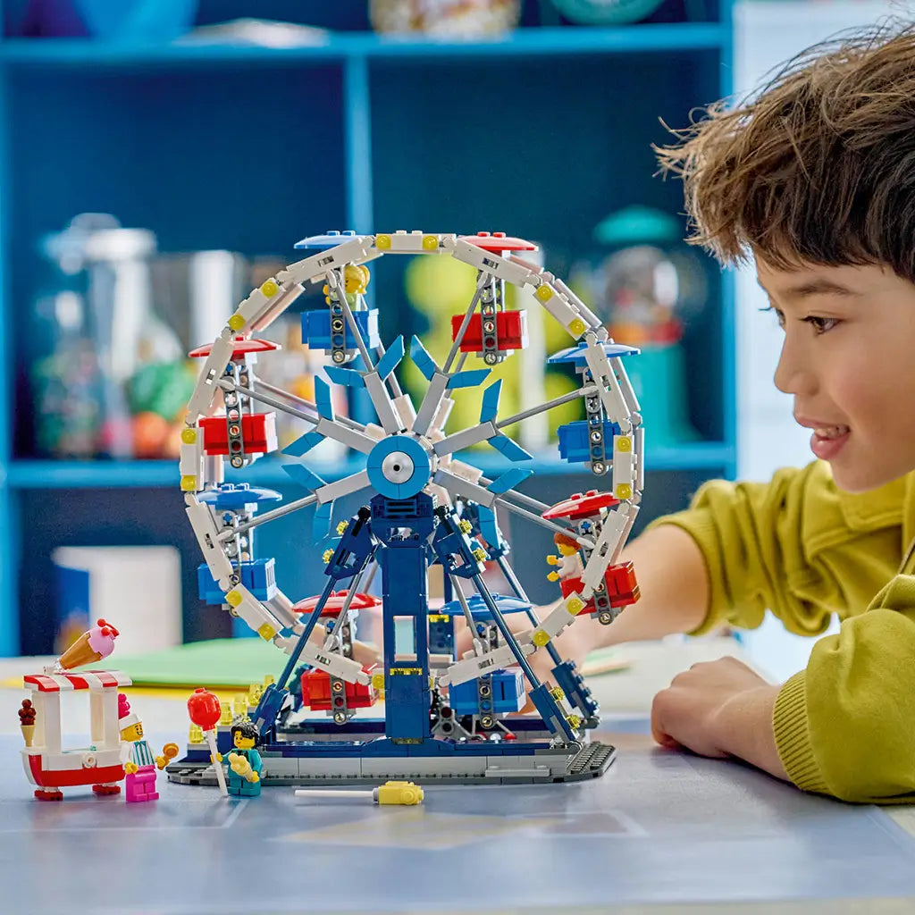 LEGO Creator Iconic Ferris Wheel in white, blue and red plastic, as a child plays beside the completed amusement ride, ticket stand and minifigures on a tabletop.