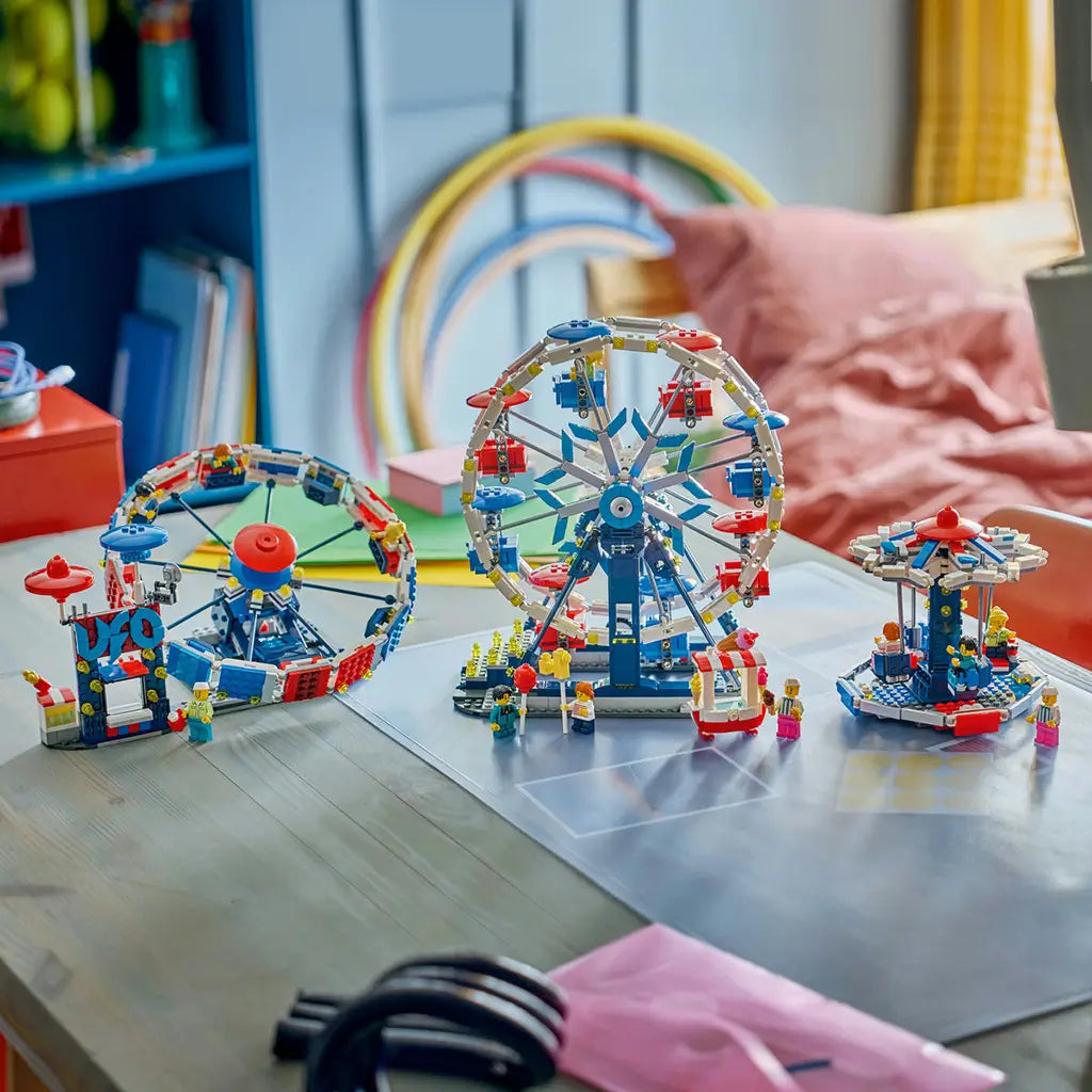 LEGO Creator amusement park rides in white, blue and red plastic, displaying a Ferris wheel, circular ride and swing ride with minifigures on a tabletop in a colorful room.
