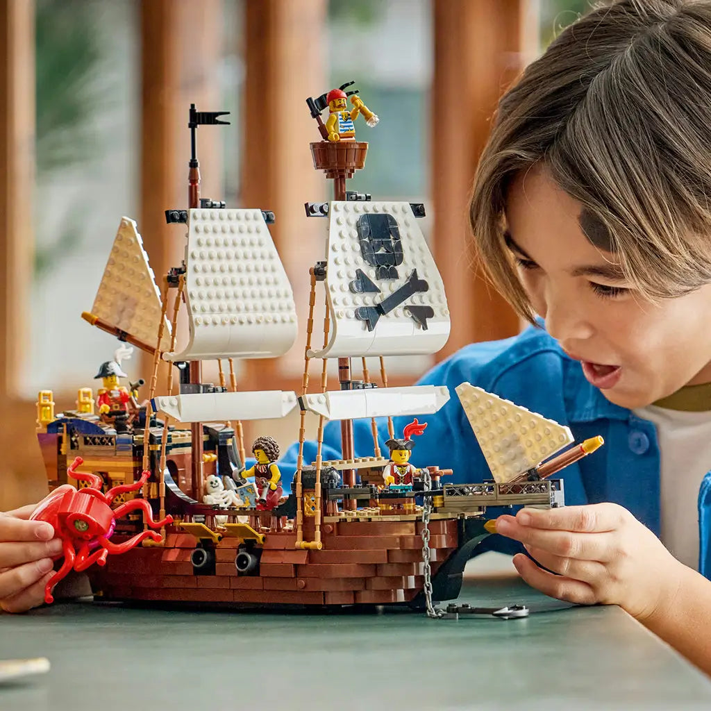 LEGO Creator Iconic Pirate Ship in brown, white and black plastic, as a child plays with the sailing ship, pirate minifigures and red sea monster on a tabletop.