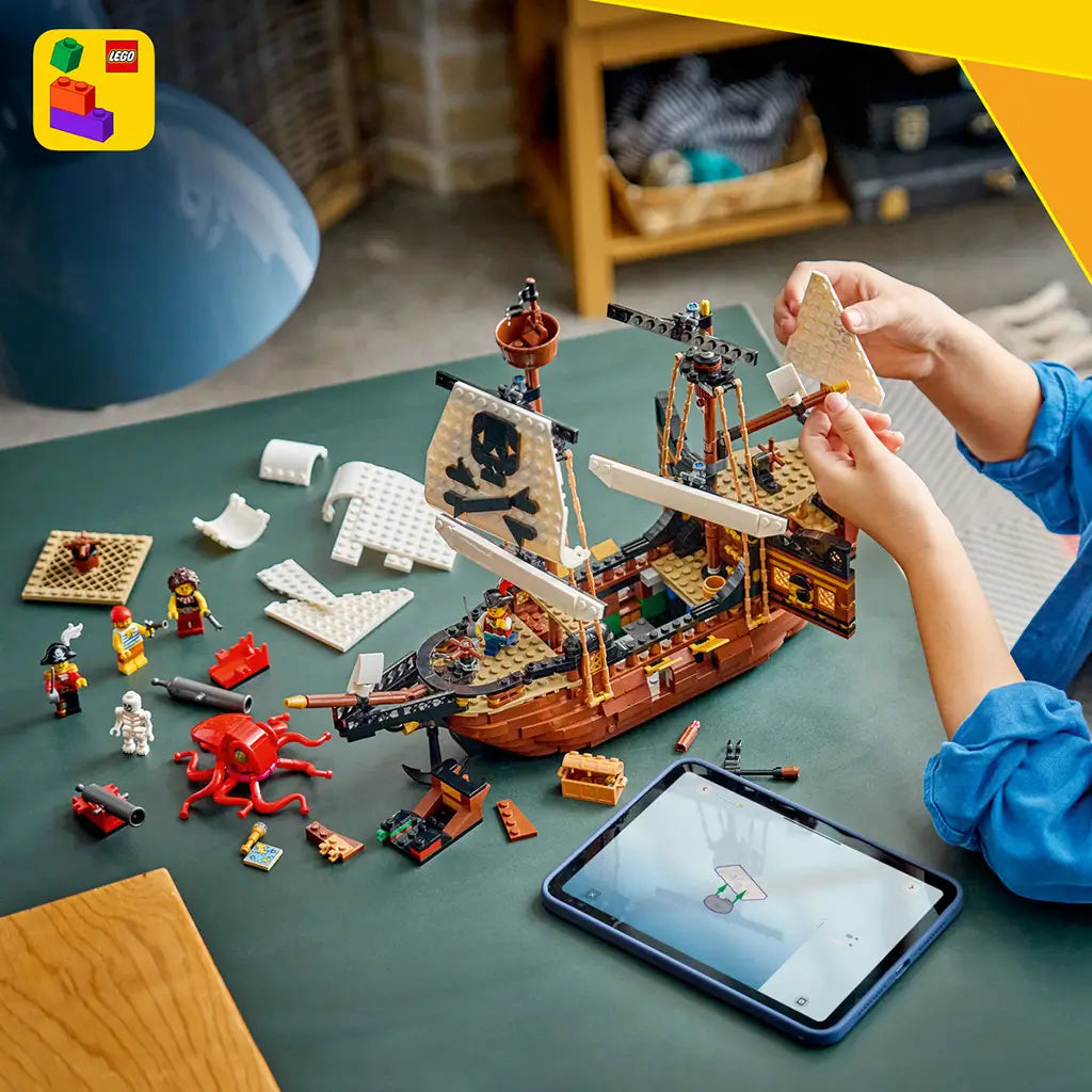 LEGO Creator pirate ship in brown, white and black plastic, as a child attaches a white sail while minifigures, a red sea monster, loose pieces and a tablet sit nearby.