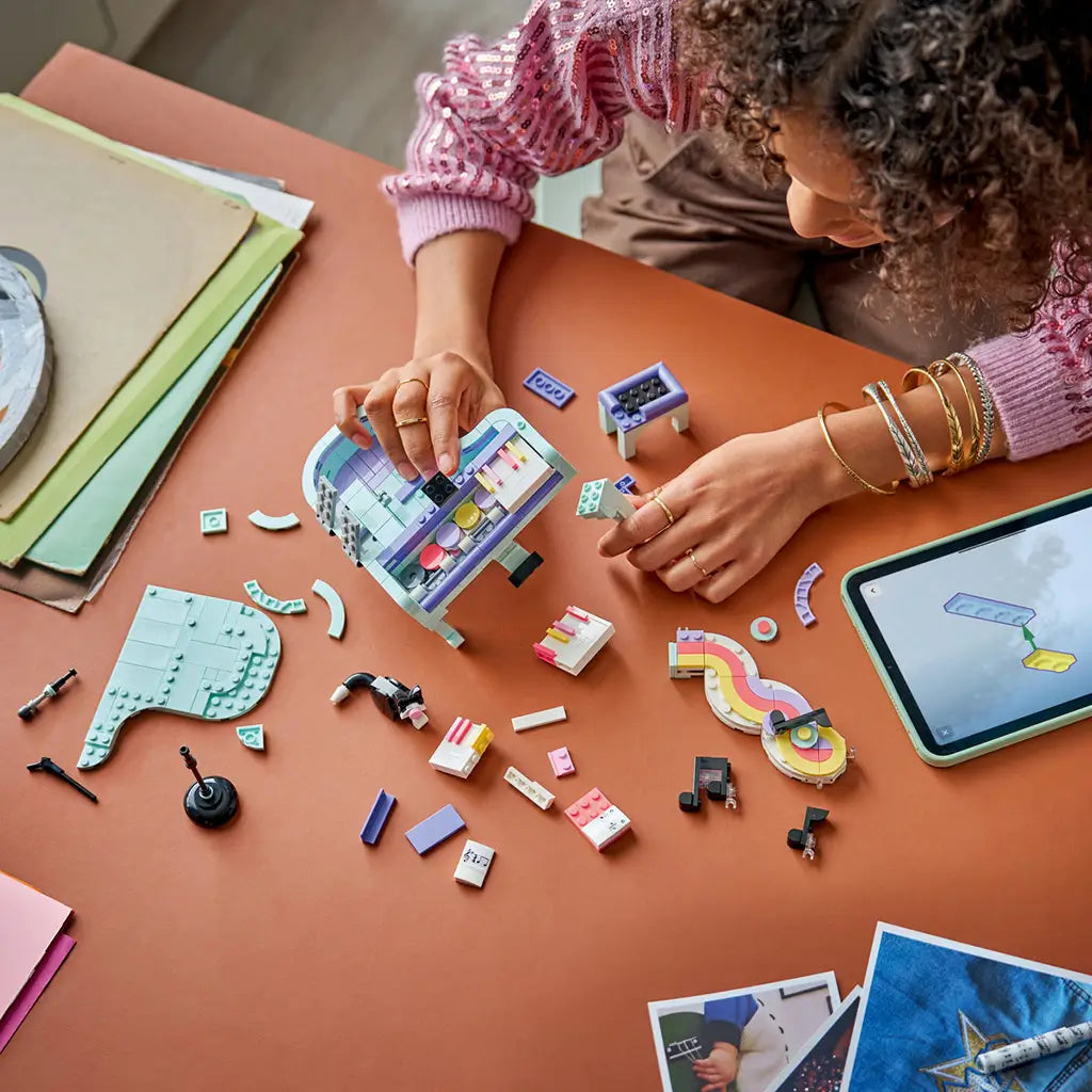 LEGO Creator Music Decor Piano set in mint, purple and pink plastic, with a person assembling the grand piano among loose bricks and digital instructions on a tabletop.