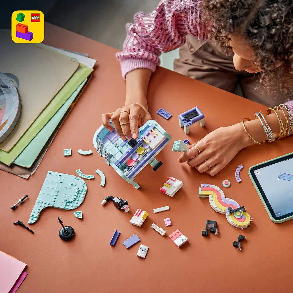LEGO Creator Music Decor Piano build in mint, purple and pink plastic, as a person assembles the piano model among colorful bricks and digital instructions on a table.