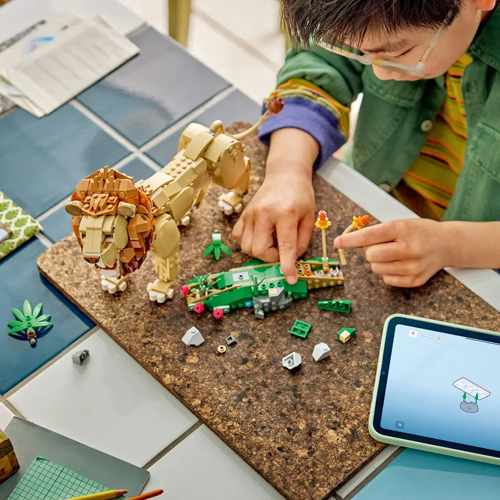 LEGO Creator Majestic Lion in tan and brown plastic stands beside green habitat pieces as a child builds plant scenery on a cork mat with a tablet nearby.