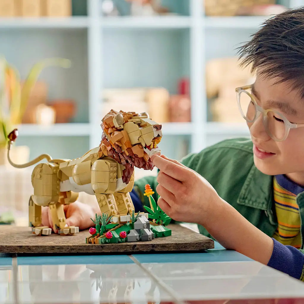LEGO Creator Majestic Lion in tan and brown plastic stands with a green flower habitat as a smiling child adjusts the lion's mane at a bright indoor craft table.