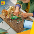 LEGO Creator Majestic Lion in tan and brown plastic stands beside a green habitat as a child assembles the plant scenery with loose bricks and a tablet nearby.