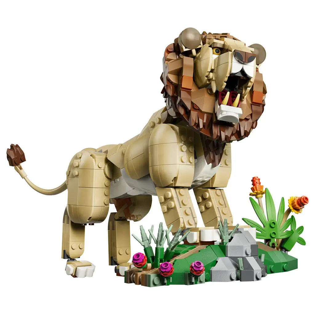 LEGO Creator Majestic Lion 31386 in tan and brown plastic, posed roaring beside a green habitat with gray rocks, grasses and colorful flowers on a white background.