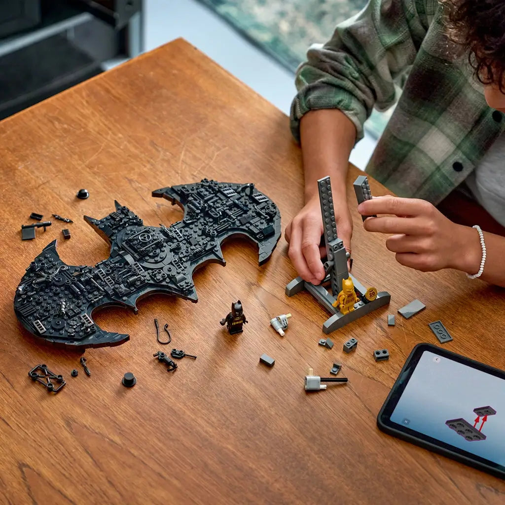 LEGO Batman logo build in black plastic pieces, complex bat-shaped form with mechanical detailing, construction toy in progress on a wooden table with scattered parts and digital guide nearby.
