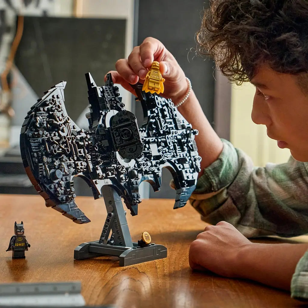 LEGO Batman logo toy in black plastic, intricate bat emblem structure on stand, display model being assembled and examined by a child at a wooden table in a bright indoor environment.