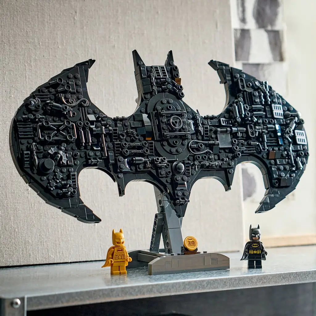LEGO Batman logo model in black plastic, detailed bat-shaped structure with layered textures, display piece positioned on a shelf with gold and classic Batman minifigures in a home setting.