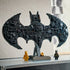 LEGO Batman logo model in black plastic, detailed bat-shaped structure with layered textures, display piece positioned on a shelf with gold and classic Batman minifigures in a home setting.