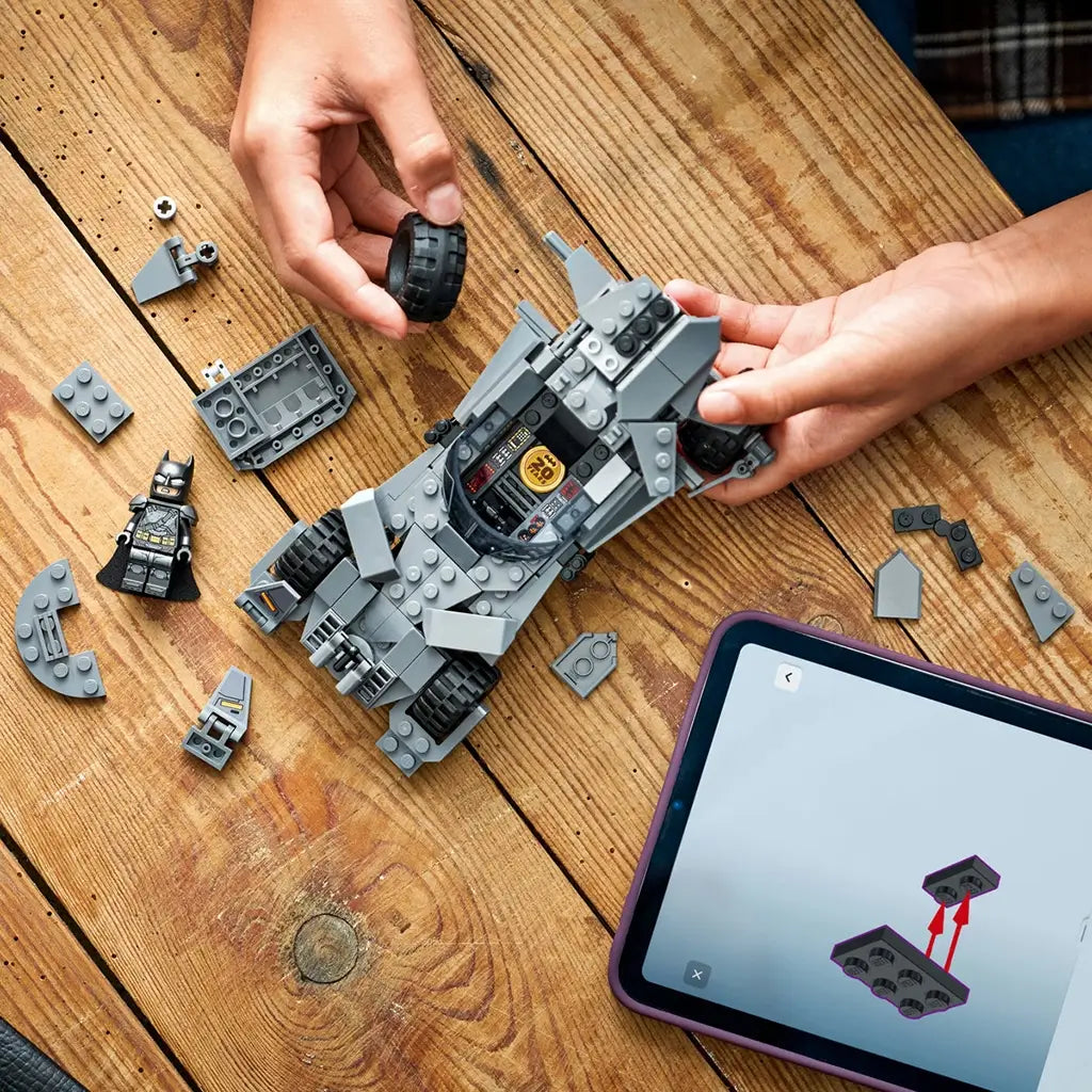 LEGO Batman Batmobile build in gray plastic pieces, angular vehicle form with cockpit and wide wheels, construction toy in progress on a wooden table with parts and digital instructions nearby.