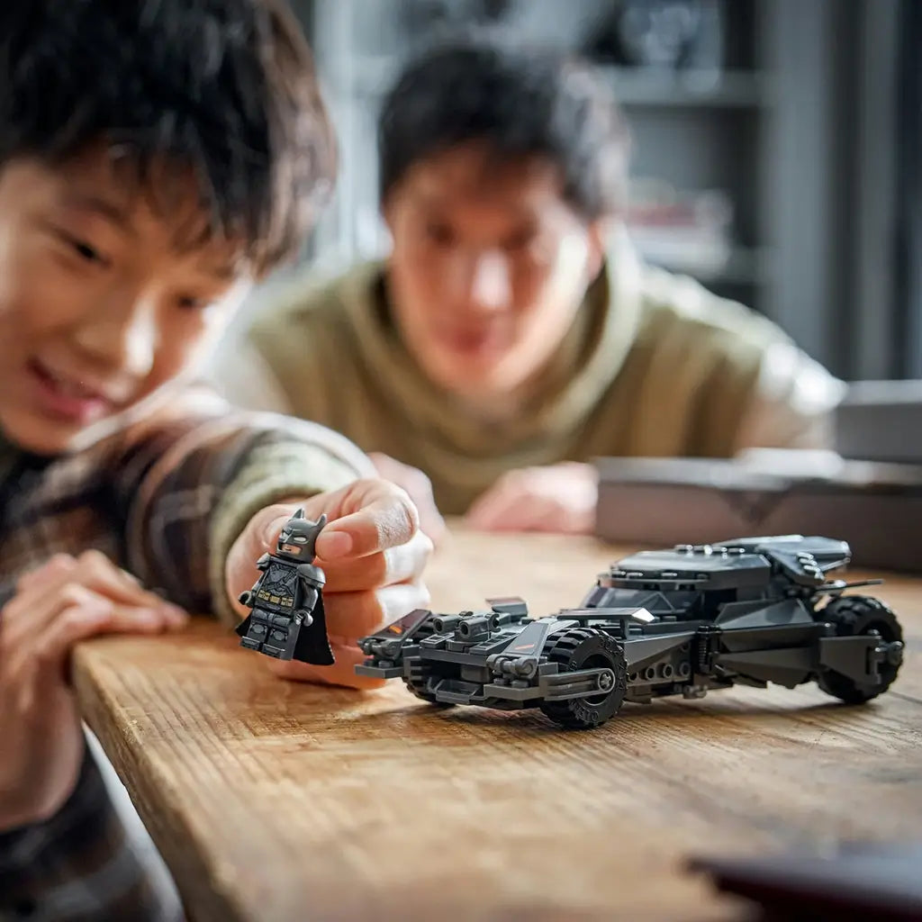 LEGO Batman Batmobile toy in gray plastic with sharp armored panels and large wheels, compact car model held and played with by a child and adult in a warm indoor home setting.