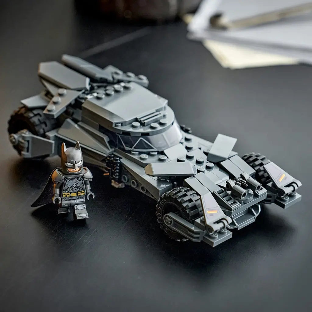 LEGO Batman Batmobile toy in dark gray plastic with angular armored body and large treaded wheels, sleek car model posed with Batman minifigure on a dark tabletop indoor setting.