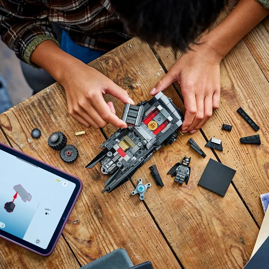 LEGO Batman Batmobile toy in black plastic bricks, angular muscle car shape, partially assembled by child hands, with loose pieces and tablet instructions on a wooden table background.