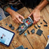 LEGO Batman Batmobile toy in black plastic bricks, angular muscle car shape, partially assembled by child hands, with loose pieces and tablet instructions on a wooden table background.