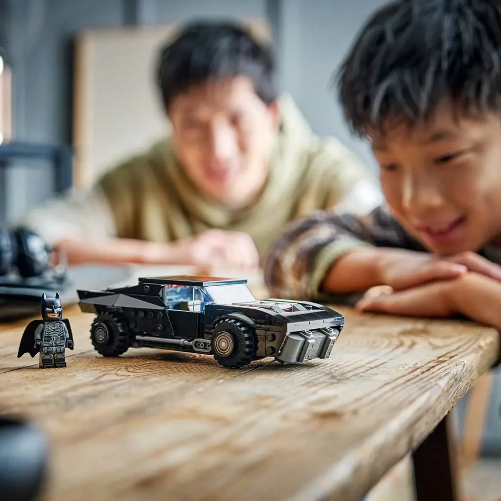 LEGO Batman Batmobile model in black plastic bricks, rugged muscle car design with large tires, displayed with Batman minifigure on a wooden table while children watch in a home setting.