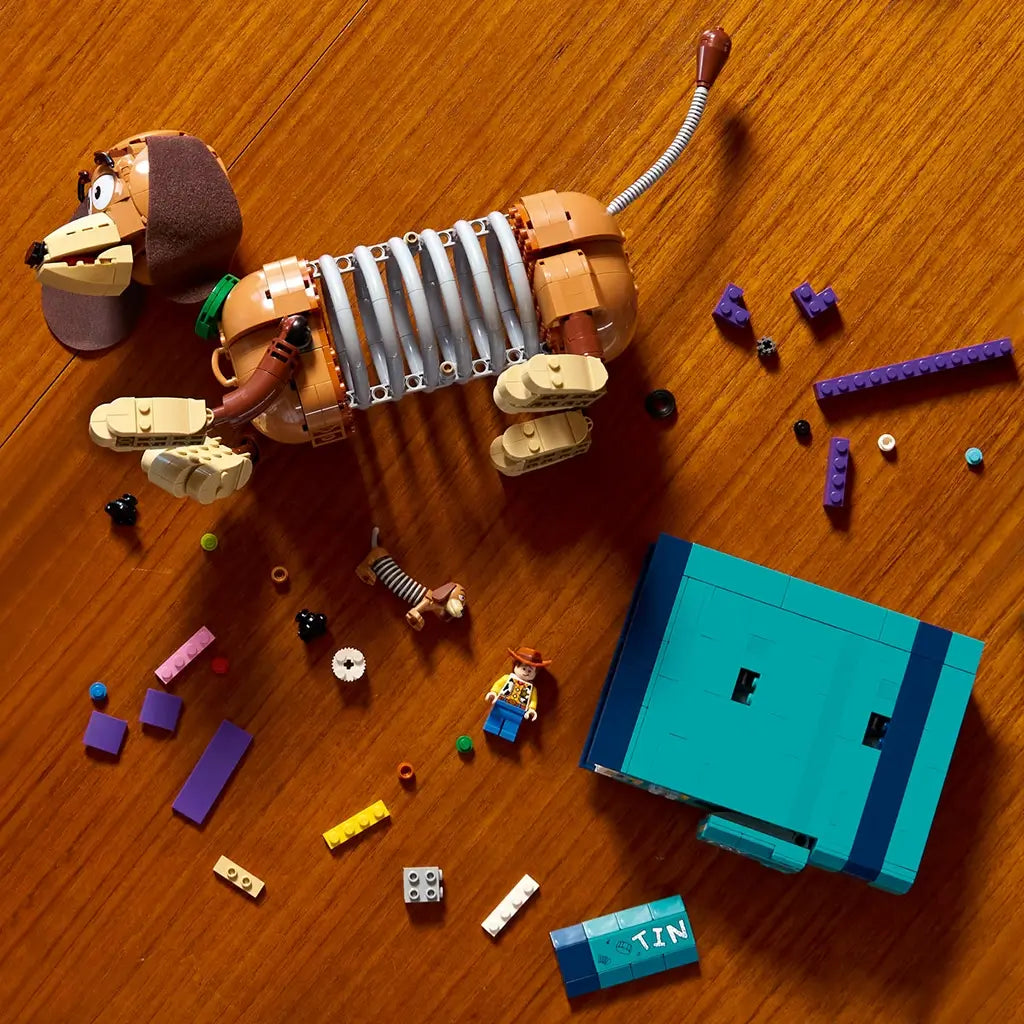 LEGO Disney Pixar Toy Story building set shows brown and tan plastic Slinky Dog sections, colorful bookend pieces, loose bricks and Woody figures arranged on a wooden tabletop during assembly.