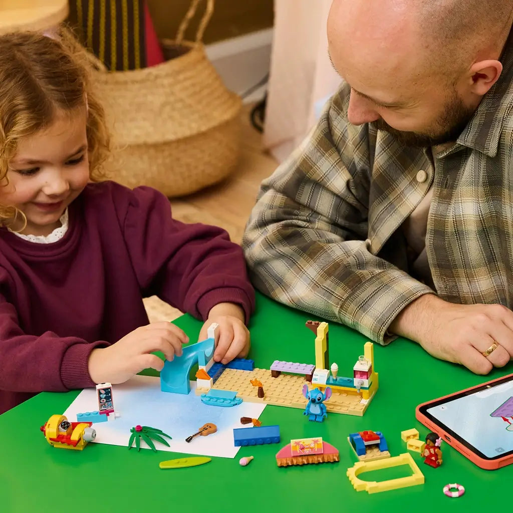 LEGO Disney Beach Fun with Lilo & Stitch set in blue, yellow, tan, and pink plastic is assembled by a child with an adult at a green table beside a tablet.
