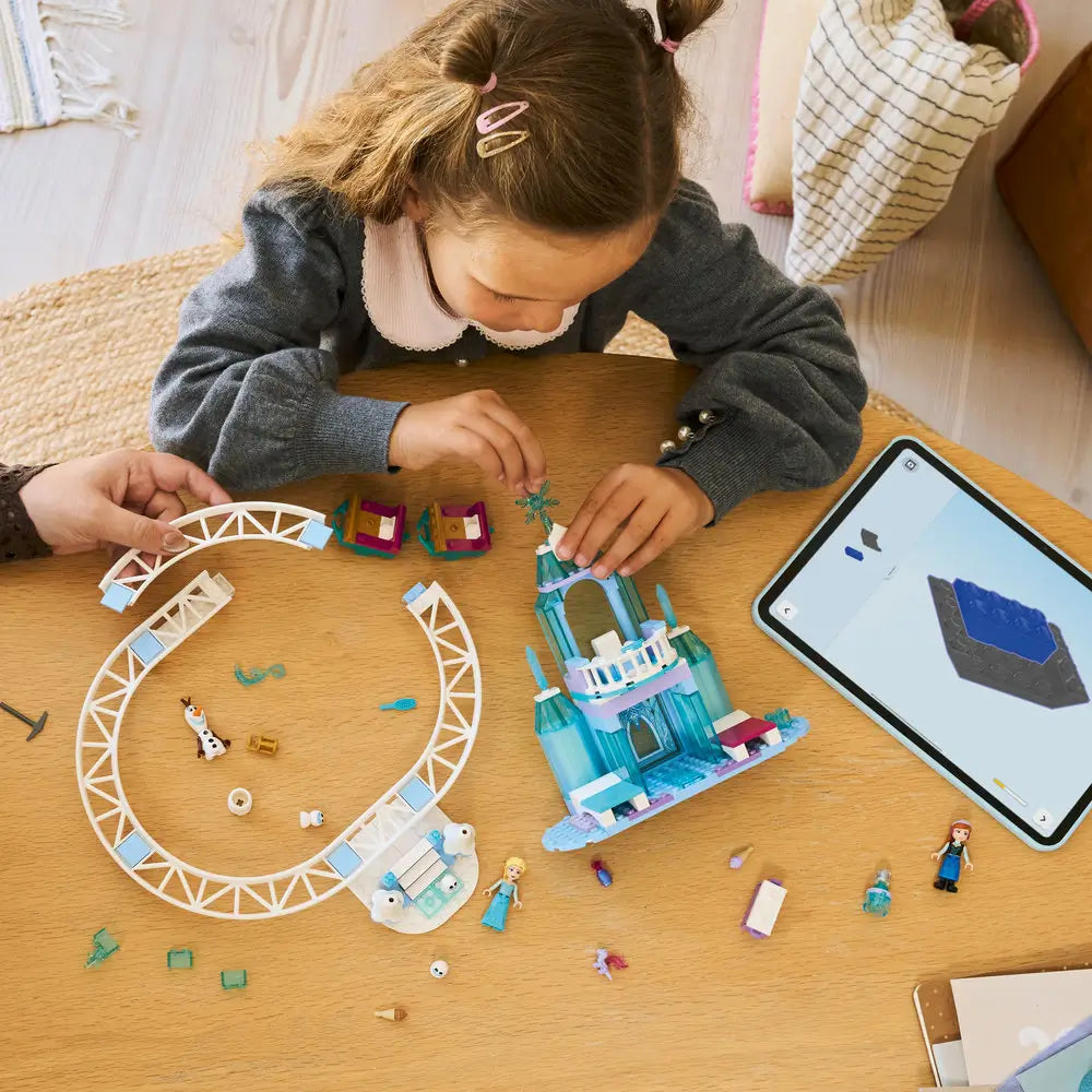 LEGO Disney Frozen building set in icy blue plastic pieces on a wooden table, partially assembled ice castle tower with curved track, showing hands-on building activity guided by a tablet app in a bright home setting.