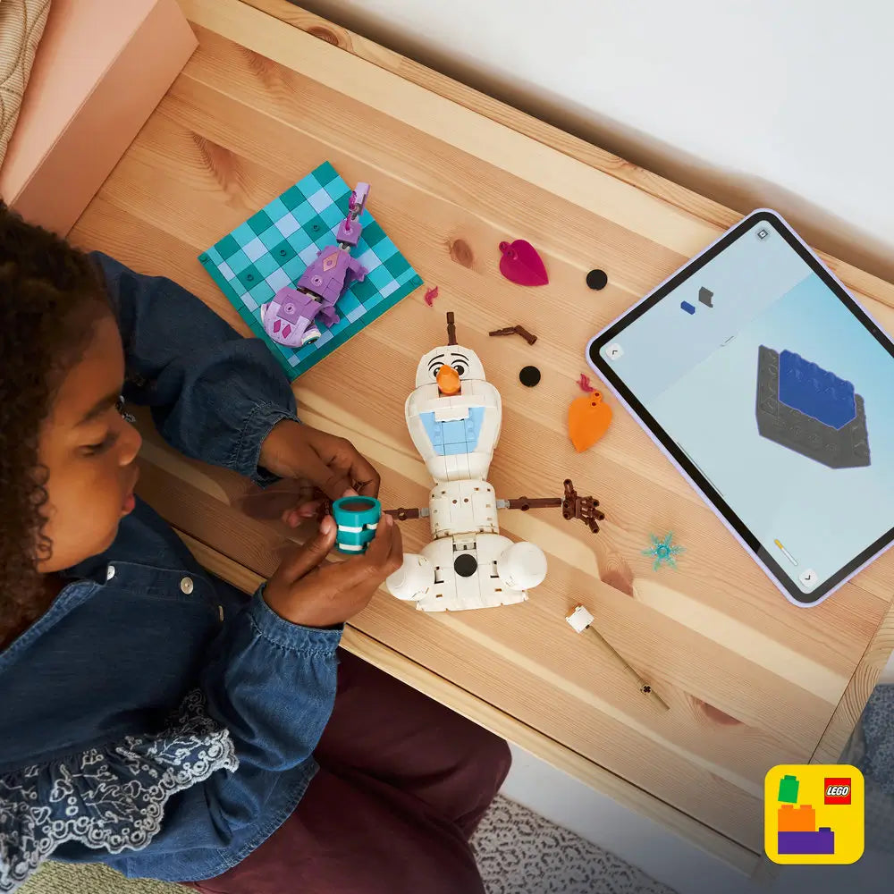 LEGO Disney Frozen Olaf playset shown during building, with white and blue plastic pieces, Bruni character, picnic elements, and a child assembling the snowman on a wooden table indoors.