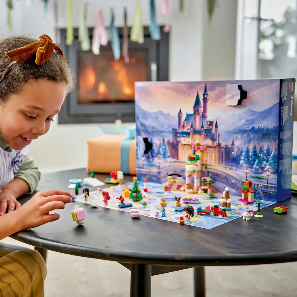 LEGO Disney Princess Advent Calendar 2026 play set shows colorful plastic figures, animals and festive builds on a snowy game mat as a child plays beside a glowing fireplace.