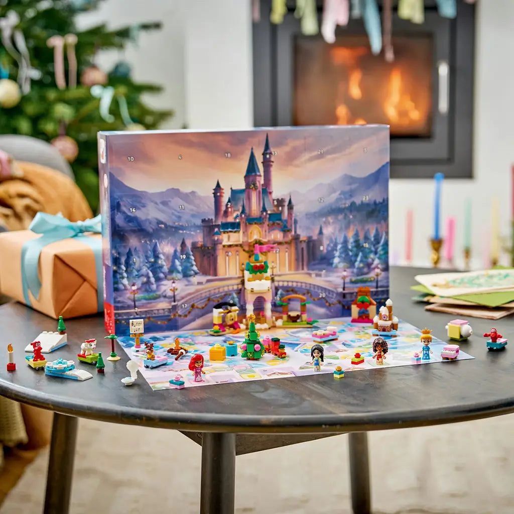 LEGO Disney Princess Advent Calendar 2026 display features colorful plastic figures, animals, a Christmas tree, train and castle tower on a snowy mat beside a glowing fireplace.