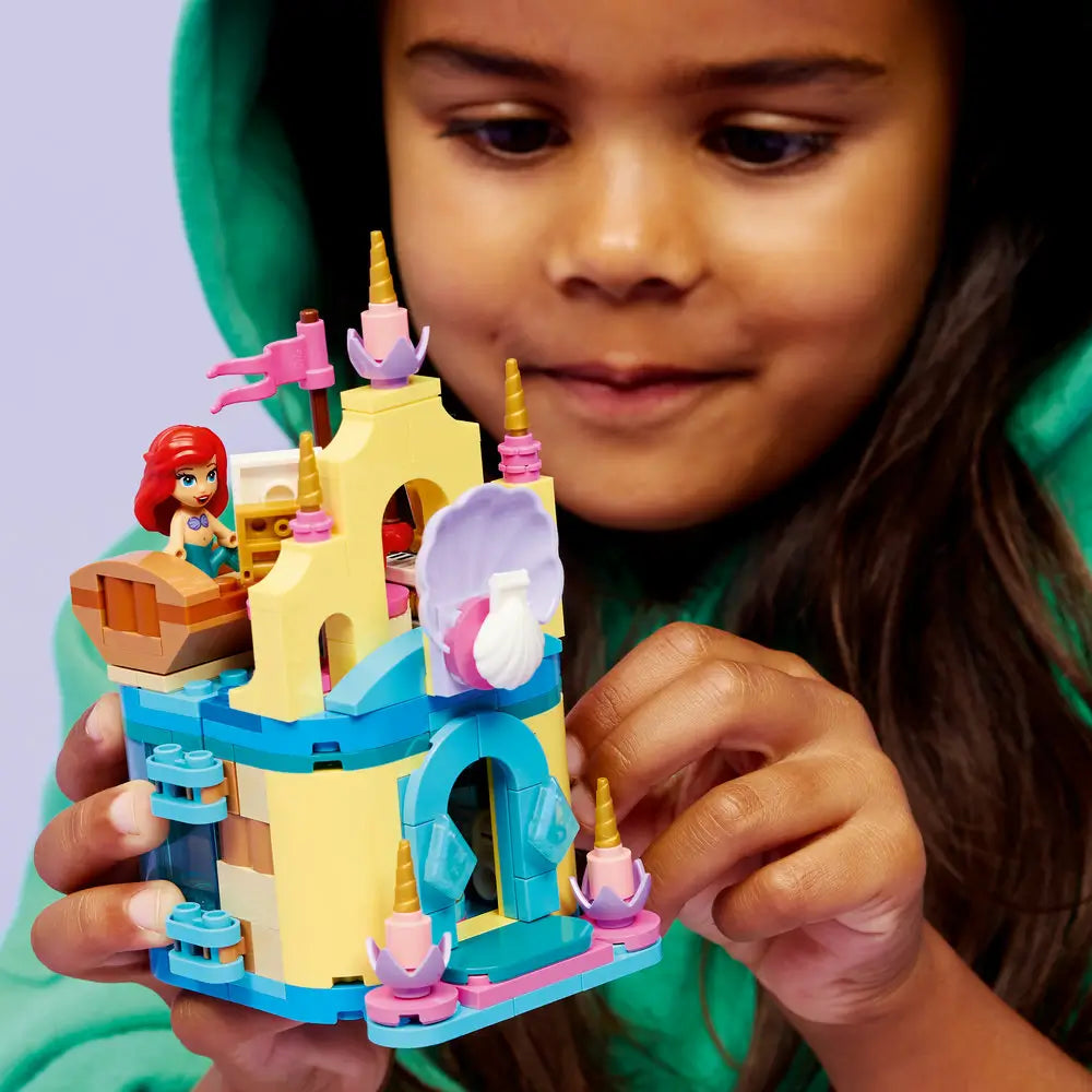 LEGO Disney Princess Ariel play scene showing a child holding the pastel mini palace with yellow towers, teal base, shell feature, and Ariel figure while smiling in a bright indoor setting.