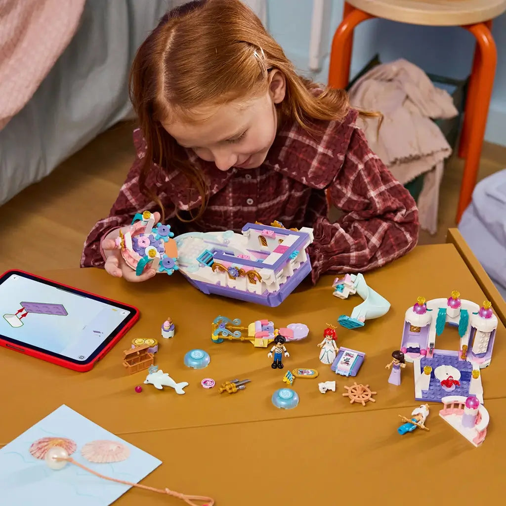 LEGO Disney Princess Ariel's Royal Wedding Boat pieces in white, lavender, pink, and teal plastic cover a table as a child assembles the modular boat beside a tablet.