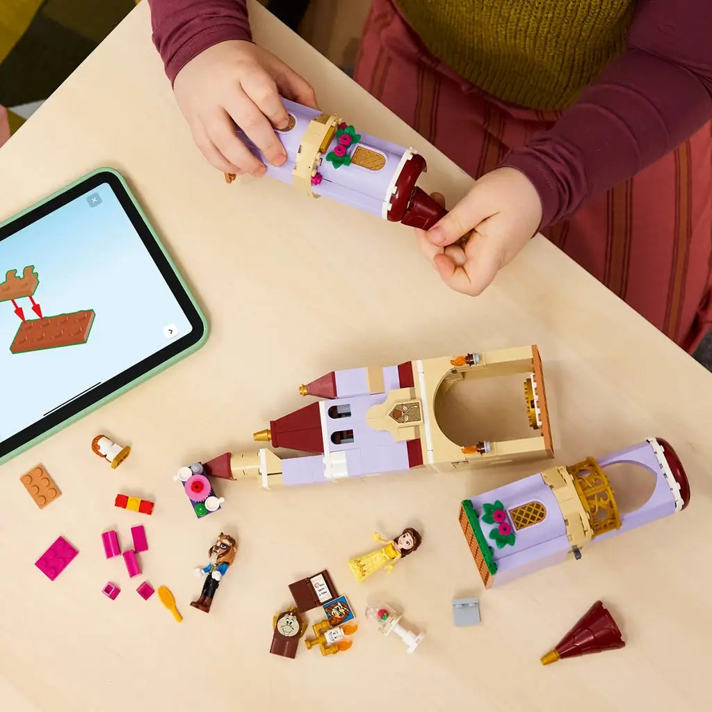 LEGO Disney Princess castle building set in lavender, tan, red, and gold plastic is assembled by a child beside tablet instructions, Belle, Beast, accessories, and tower pieces.
