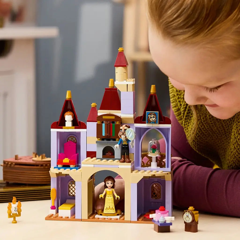LEGO Disney Princess castle in lavender, tan, red, and gold plastic is explored by a child at a table, with Belle, Beast, Cogsworth, Lumiere, rose, and furnished rooms.