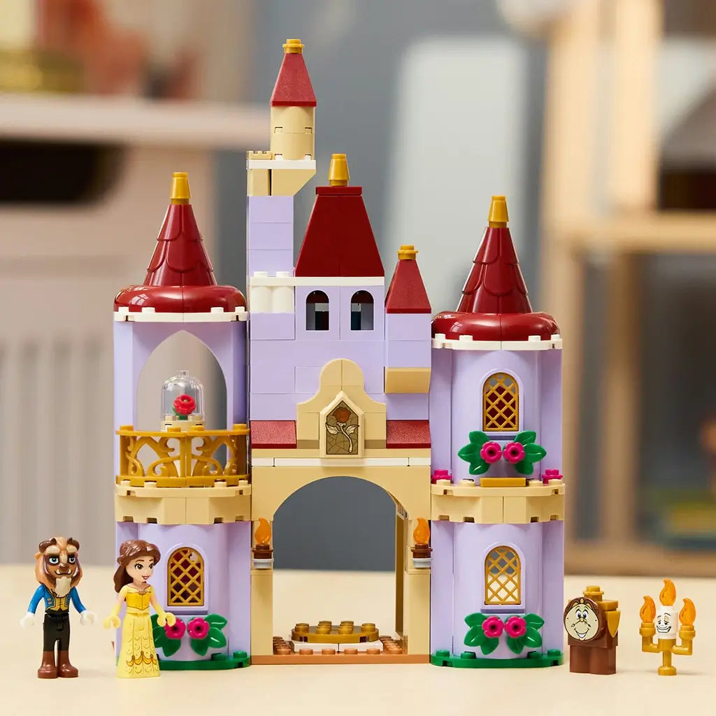 LEGO Disney Princess castle set in lavender, tan, red, and gold plastic shows Belle and Beast beside an arched castle with rose balcony, flowers, Lumiere, and Cogsworth.