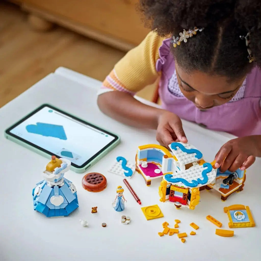 LEGO Disney Princess Cinderella dress set in blue, white and gold plastic with open wardrobe sections and build pieces as a child follows digital instructions on a tablet at a white table indoors.