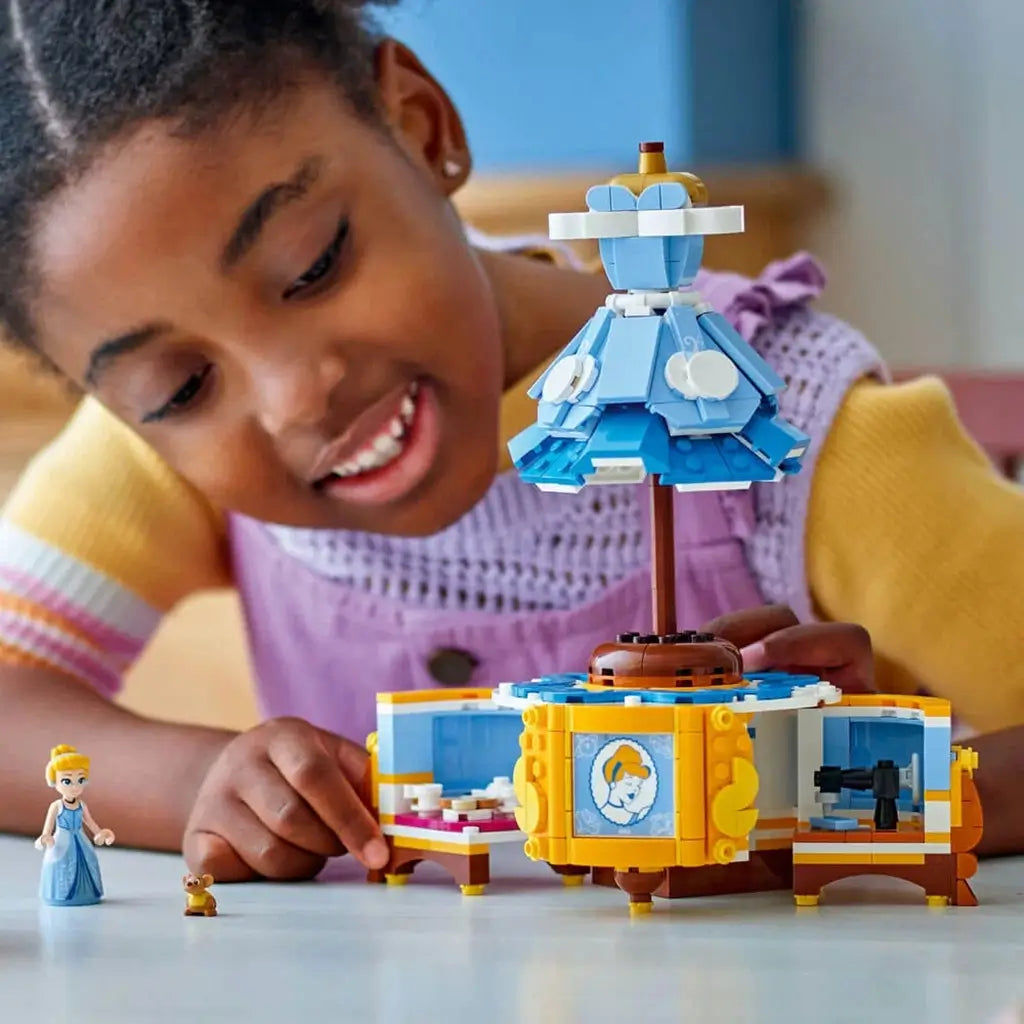 LEGO Disney Princess Cinderella dress model in blue, white and gold plastic with opening display cabinet as a smiling child explores the build on a tabletop in a bright indoor setting.