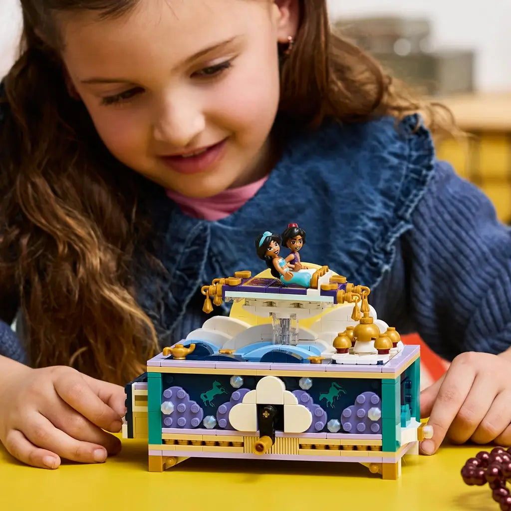 LEGO Disney Princess Jasmine's Jewelry Box in teal, lavender, white, and gold plastic sits on a yellow table as a child plays with the finished crank box and character figures.