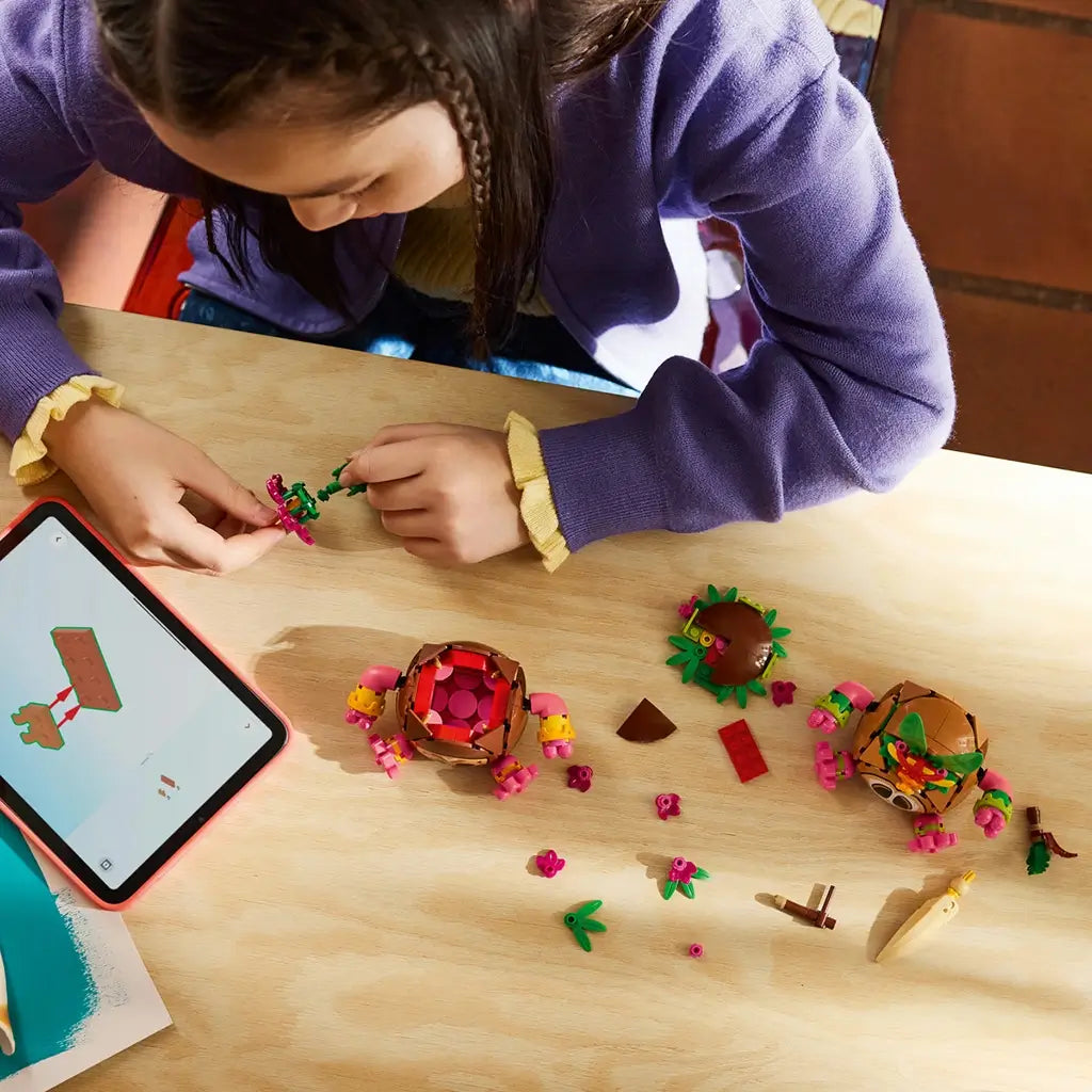 LEGO Disney Moana Kakamora building set in brown, pink, green, and tan plastic is assembled by a child on a wood table with loose tropical pieces and tablet instructions nearby.