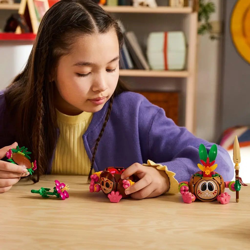 LEGO Disney Moana Kakamora figures in brown, pink, green, and tan plastic are played with by a child at a wood table, with floral headpieces, flower accessory, and spear.