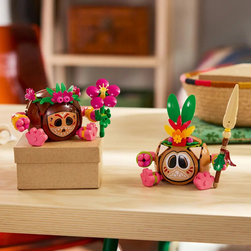 LEGO Disney Moana Kakamora figures in brown, pink, green, and tan plastic are displayed on a wood table, one holding a purple flower and the other a tall tan spear.