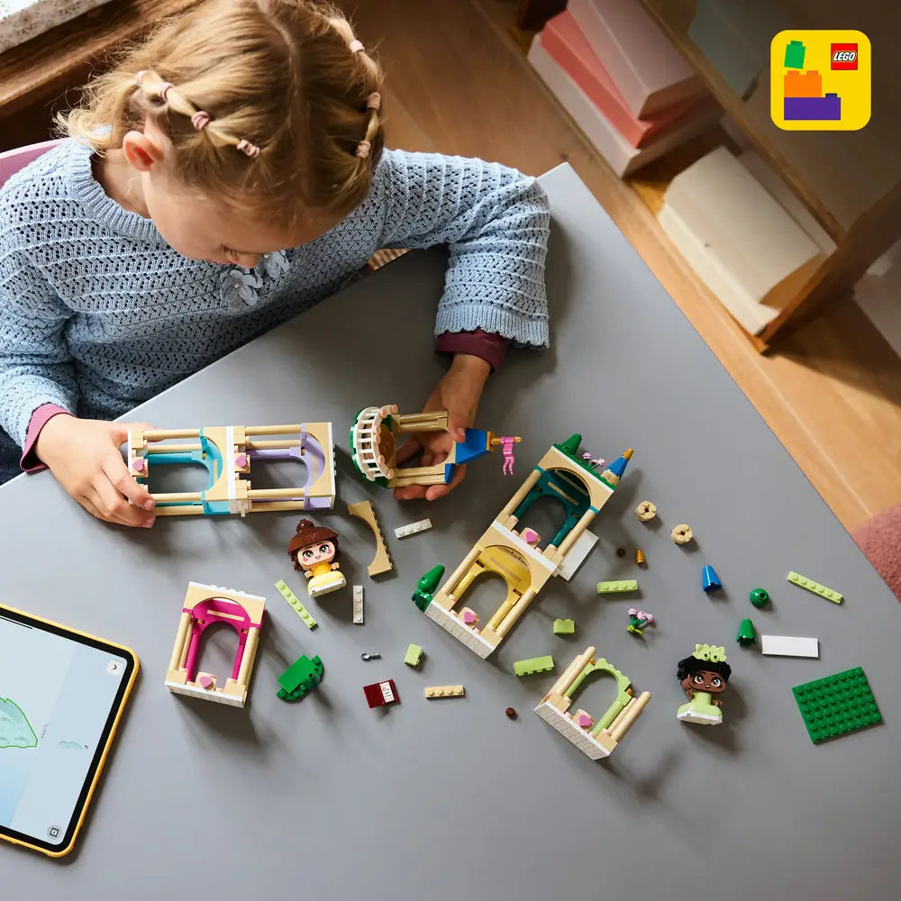 LEGO Disney Princess building toy shown in use, with pastel tan arches, blue roof elements, and character figures spread across a table as a child assembles the castle in a bright indoor setting.