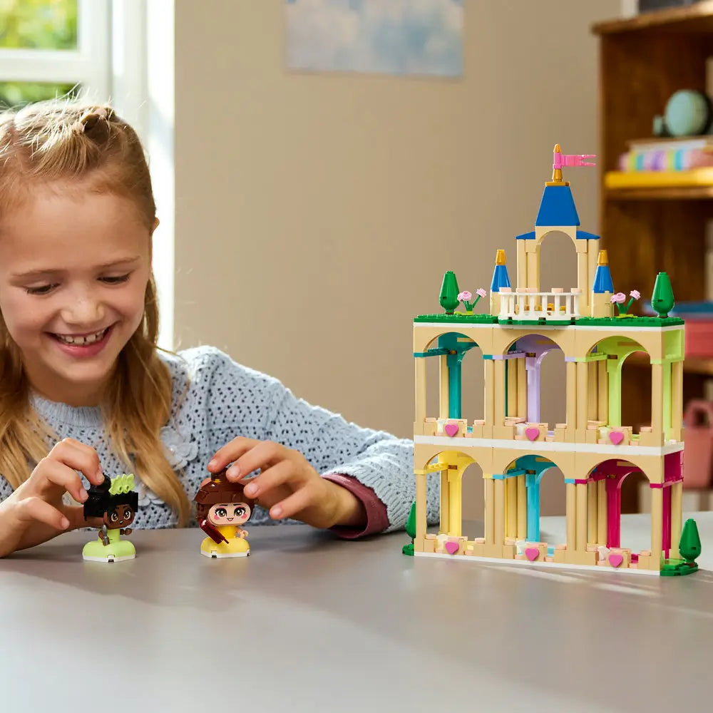 LEGO Disney Princess building experience showing a child holding Belle and Tiana mini figures beside a completed pastel castle with tan arches, blue towers, and heart decorations indoors.